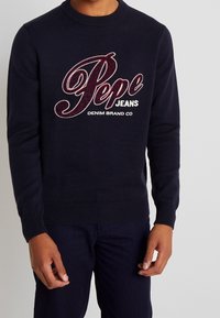 Maglione lavorato a maglia blu navy con polsini e orlo a costine, caratterizzato da un grande logo ricamato in rosso "Pepe Jeans" sul davanti. Dettaglio del marchio in denim incluso.