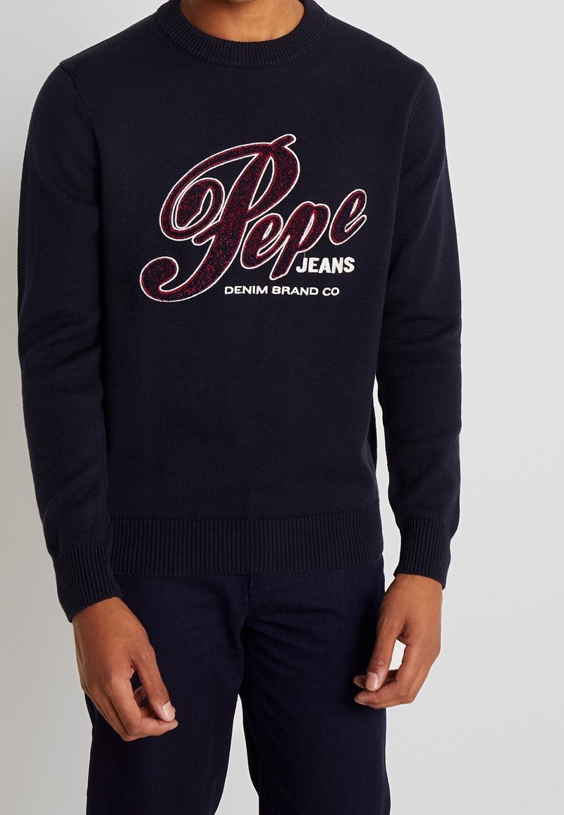 Maglione lavorato a maglia blu navy con polsini e orlo a costine, caratterizzato da un grande logo ricamato in rosso "Pepe Jeans" sul davanti. Dettaglio del marchio in denim incluso.