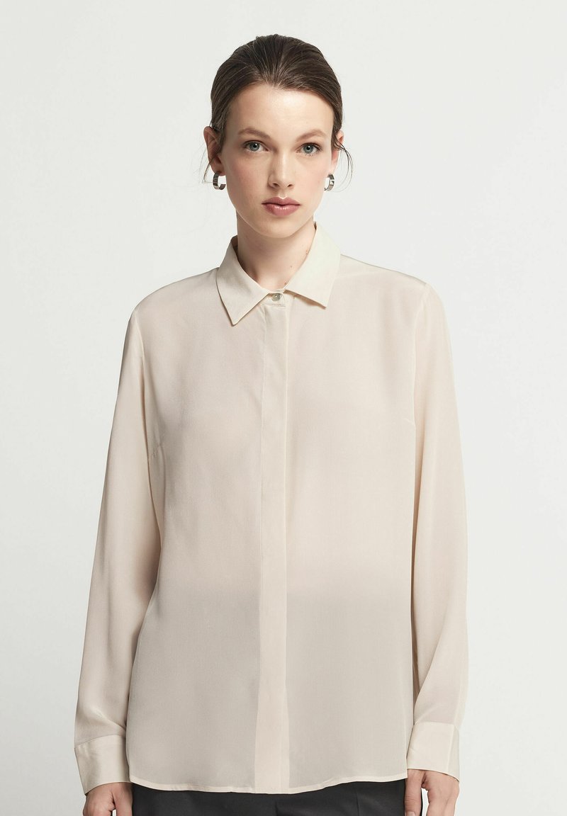 Ljusbeige långärmad blus med spetsig krage, knäppning fram och slät, genomskinlig tygtextur. Skräddarsydd passform med manschettärmar.