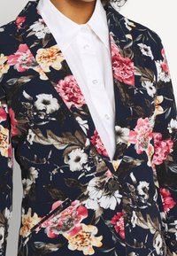 Saka s květinovým vzorem v námořnické barvě, s růžovými, bílými a oranžovými květy. Má klopový límec a černý knoflík, pod ním bílou košili.