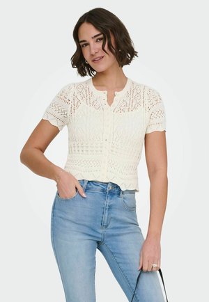 Kobieta o krótkich, ciemnych włosach, ubrana w białą koronkową bluzkę na krótki rękaw z guzikami oraz jasnoniebieskie dżinsy z wysokim stanem, trzymająca czarny pasek.