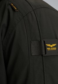 Giacca nera in tessuto con texture a coste. Presenta un'etichetta con dettagli gialli e una fascia nera con fibbia vicino alla cucitura della spalla.