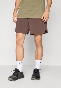 Calções atléticos castanhos feitos de tecido leve, com um pequeno logótipo da Nike. Usados com ténis pretos e meias brancas de cano curto.