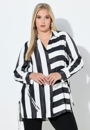 Vaaleahiuksinen nainen, jolla on suuret renkaanmuotoiset korvakorut ja mustavalkoraidallinen pitkähihainen pusero, jossa on kiristysnyörit, poseeraa valkoista taustaa vasten.