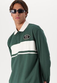 Mladý muž s tmavými slunečními brýlemi a zeleným ragbyovým tričkem s bílým límcem a nášivkou "Prosím, myslete" na hrudi.