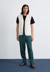 En ung mand står foran en ensfarvet baggrund, iført en hvid veste over en sort t-shirt og mønstrede grønne bukser, der suppleres af mørke sneakers.