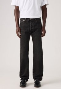 Μαύρο τζιν από denim με άνετη εφαρμογή, με πέντε τσέπες, χαλκινούς δακτυλίους και κλείσιμο με κουμπί. Συνδυασμένο με λευκό t-shirt και μαύρα παπούτσια.