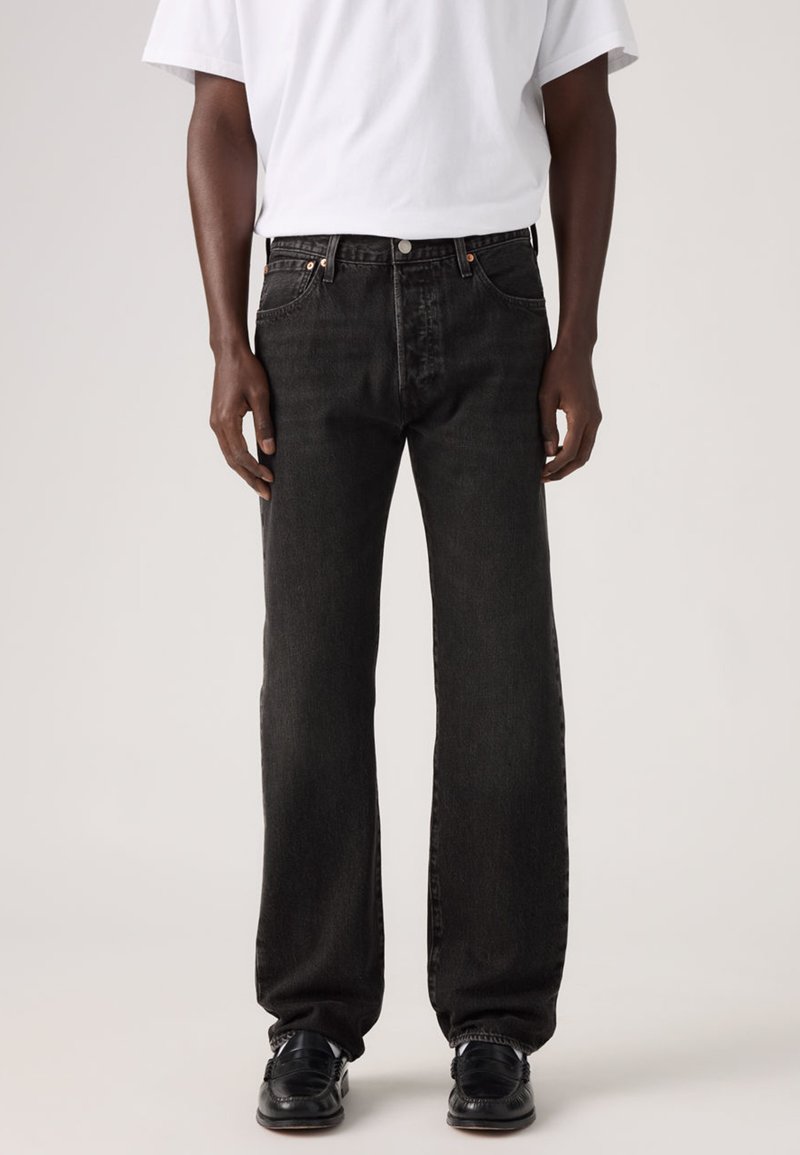 Μαύρο τζιν από denim με άνετη εφαρμογή, με πέντε τσέπες, χαλκινούς δακτυλίους και κλείσιμο με κουμπί. Συνδυασμένο με λευκό t-shirt και μαύρα παπούτσια.