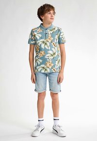 Lichtblauw poloshirt met korte mouwen en bloemenpatroon, met groene bladeren en gele bloemen. Gecombineerd met opgerolde denim shorts en witte sneakers.