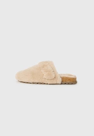 Sabots en peluche beige à enfiler avec une semelle en liège et une boucle décorative sur le côté, présentés de profil sur un fond uni.
