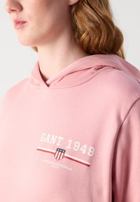 Sudadera rosa hecha de tela suave con un gráfico frontal que dice "GANT 1949" y un diseño de escudo, con capucha ajustable y corte holgado.