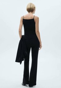 Černý bez rukávů top v kombinaci s černými kalhotami, ukazující přiléhavý design. Celý vzhled doplňuje černý blazer přehozený přes ruku.