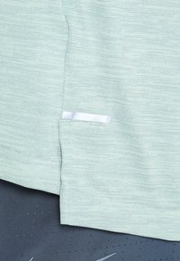 Lys grøn tekstureret stoftop med et lille hvidt mærke ved falden, placeret på en grå, perforeret stofbaggrund.