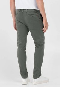 Pantalons en coton vert, ajustés, avec deux poches arrière et une fermeture éclair. Portés avec des baskets blanches, mettant en valeur une texture lisse.