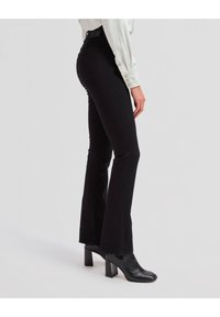 Femme portant un pantalon noir évasé ajusté, des bottines noires à talons et une chemise blanche à manches longues, debout sur un fond clair uni.