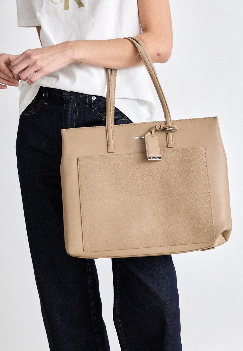 Personne portant un t-shirt blanc et un jean foncé tenant un grand sac fourre-tout beige Calvin Klein avec une poche avant et des poignées doubles.