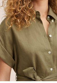 Olivengrøn linnedskjorte, korte ærmer, knaplukning foran og bindedetalje ved taljen. Glat tekstur med en subtil glans.