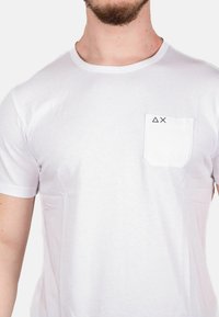 T-shirt bianco a maniche corte realizzato in cotone, con una tasca sul petto decorata da cuciture geometriche nere. Texture liscia, design semplice.