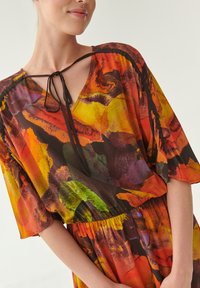 Kleurvolle blouse met een v-hals, strikdetail bij de halslijn en korte, gerimpelde mouwen. De stof vertoont een levendig abstract patroon in rood, oranje en groen.