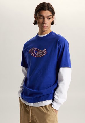 Joven con camiseta azul de Vans en capas sobre mangas largas blancas y pantalones beige, mirando hacia abajo y de lado contra un fondo liso.