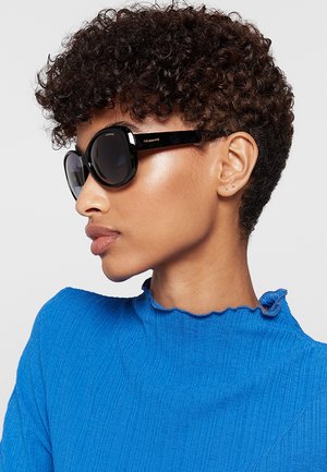 Persona con capelli corti e ricci indossa grandi occhiali da sole neri e una maglia a coste blu brillante con scollo arricciato, rivolta verso sinistra.