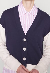 Marinblå och gräddfärgad v-ringad kofta med långa ärmar, fem knappar och ribbade muddar. Under finns en rosa och vit randig skjorta.