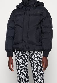 Svart puffjacka med huva, med en kviltad design och elastiska ärmslut, tillsammans med svarta byxor prydda med vita blommönster.