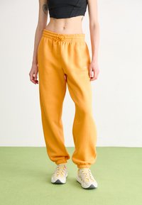 Persona que lleva pantalones de chándal naranja brillante, zapatillas blancas y amarillas, y un top corto negro, de pie sobre un suelo verde claro contra un fondo liso.