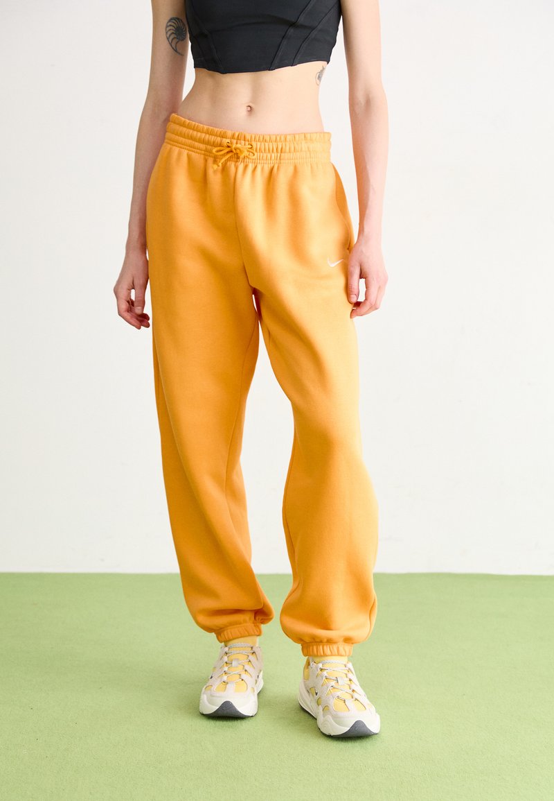 Persona que lleva pantalones de chándal naranja brillante, zapatillas blancas y amarillas, y un top corto negro, de pie sobre un suelo verde claro contra un fondo liso.