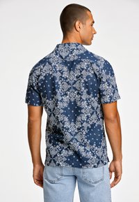 Camisa de manga corta de color azul marino con un patrón de cachemira y puntos blancos, con cuello clásico y dobladillo recto. Usada con unos vaqueros de color azul claro.