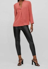 Blusa di colore corallo con texture, scollo a V e maniche a tre quarti, abbinata a leggings neri lucidi e tacchi neri con cinturini.