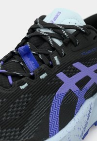 Μαύρο αθλητικό παπούτσι με υφασμάτινο επάνω μέρος, μοβ λεπτομέρειες και ελαφριά σόλα με πιτσιλιές. Τα κορδόνια είναι μαύρα και λευκά.