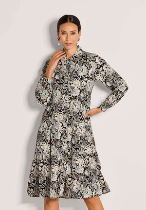 Frau in einem langärmligen, knielangen Blumenmusterkleid mit Kragen und Knopfdetails, die mit einer Hand nahe dem Gesicht und der anderen auf der Hüfte posiert.