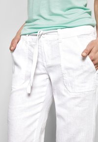 Pantalons en lin blanc avec une taille élastique et un cordon de serrage, dotés de deux poches avant de forme carrée, sur fond clair.