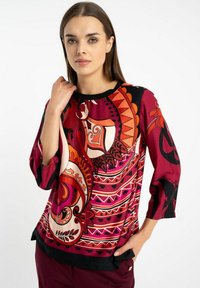 Wielokolorowa bluzka z odważnymi wzorami paisley w kolorach czerwonym, pomarańczowym i czarnym, uszyta z lekkiego materiału, z rękawami o długości trzech czwartych i okrągłym dekoltem.