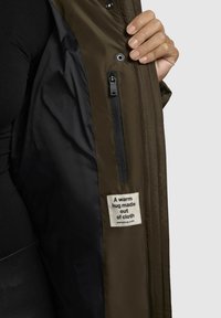 Bruine geïsoleerde jas met een zwarte voering, een ritszak en een stoffen etiket met de tekst "Een warme knuffel van stof."