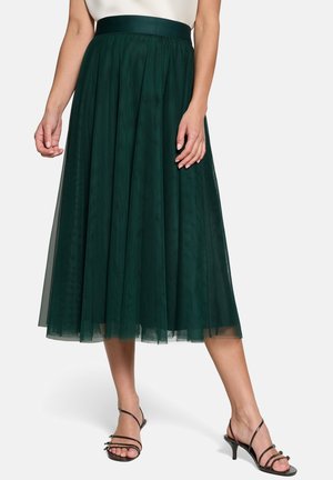 Femme portant une jupe midi en tulle vert foncé, un haut blanc et des sandales à talons noires à brides, debout devant un fond uni.