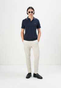 Mornarska polo majica s bijelim obrubom, bež hlače i crne loafers cipele. Model nosi sunčane naočale. Minimalistički dizajn; jednostavan kroj.