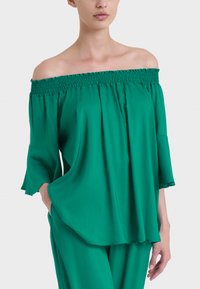 Top verde off-the-shoulder con scollo smock, vestibilità ampia e maniche a campana. Realizzato in tessuto morbido, presenta un design fluido.