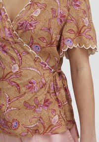 Blusa avvolgente in tessuto marrone con motivi floreali rosa, maniche scallop e dettaglio a fiocco in vita. Tessuto morbido, vestibilità comoda.