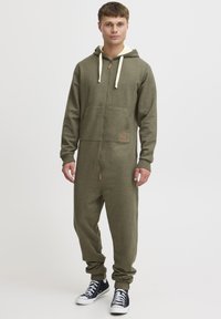 Olive green zip-up jumpsuit with a hood, cream drawstrings, front pockets, and ribbed cuffs. Worn with black sneakers.