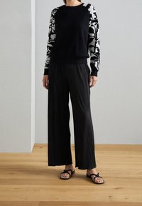 Svart tröja med blommönstrade ärmar, matchad med vida, plisserade svarta byxor. Skorna är platta, svarta flip-flop-sandaler.