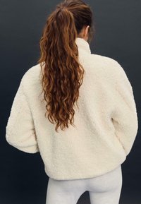 Bela puhasta flis jakna z visokim ovratnikom in sproščenim krojem, s super mehko teksturo in spuščenimi rameni, gledano s hrbta.