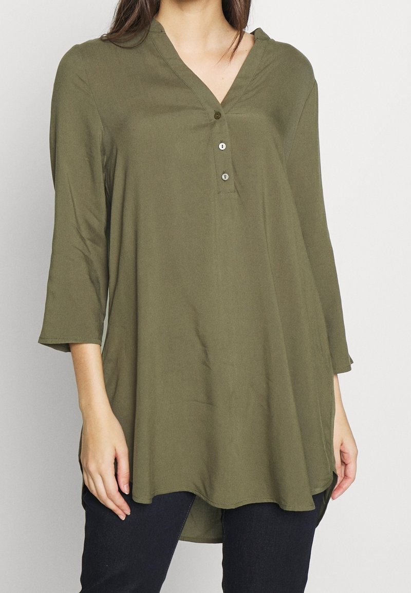 Tunique vert olive avec un décolleté en V, trois boutons sur le devant et des manches mi-longues. Présente un ourlet arrondi et un tissu doux.