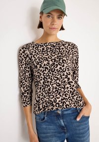 Donna con un cappello verde, una maglietta a maniche lunghe con stampa leopardata beige e nera, e jeans blu, con una mano in tasca su uno sfondo bianco.