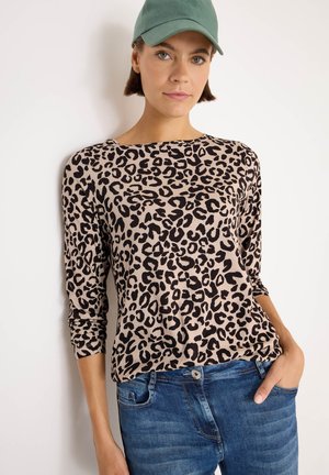 Kvinna som bär en grön mössa, en beige och svart leopardmönstrad långärmad topp och blå jeans med en hand i fickan mot vit bakgrund.