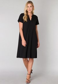Zwarte knielange jurk met V-hals en korte mouwen. Gemaakt van zachte stof en voorzien van zijopeningen voor meer bewegingsvrijheid.