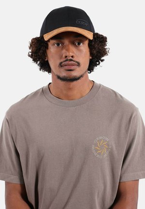 Homme aux cheveux bouclés portant une casquette Oxbow noire et marron ainsi qu'un t-shirt taupe Oxbow avec un logo soleil jaune sur la poitrine, sur un fond blanc uni.