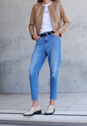 Personne portant une veste courte beige, un t-shirt blanc, un jean bleu taille haute avec une ceinture noire et des mocassins crème, debout à l'intérieur.