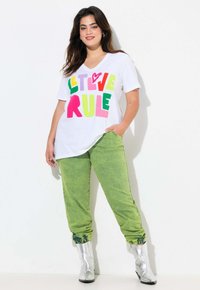 Camiseta blanca de cuello en V con el texto colorido "QUE EL AMOR GOBIERNE". Combinada con pantalones verdes brillantes y botas de tobillo plateadas, mostrando un ajuste relajado.